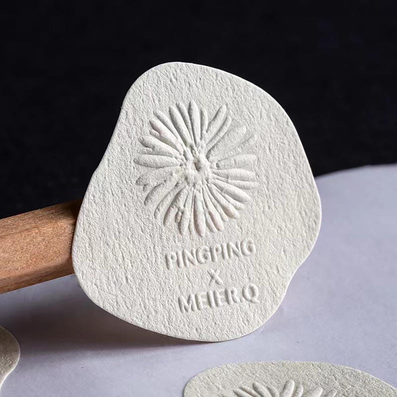 Puede incluir: Etiqueta de papel texturizado blanquecino con un dise&ntilde;o floral en relieve. La etiqueta tiene una forma irregular y presenta el texto "PINGPING X MEIERQ" en una fuente sans-serif. La etiqueta oscurece parcialmente un l&aacute;piz de madera.