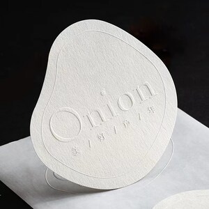 Puede incluir: Una tarjeta de papel blanco texturizado con la palabra "Onion" en relieve en el centro. La tarjeta tiene una forma org&aacute;nica &uacute;nica con bordes redondeados y elementos de dise&ntilde;o y texto en relieve.