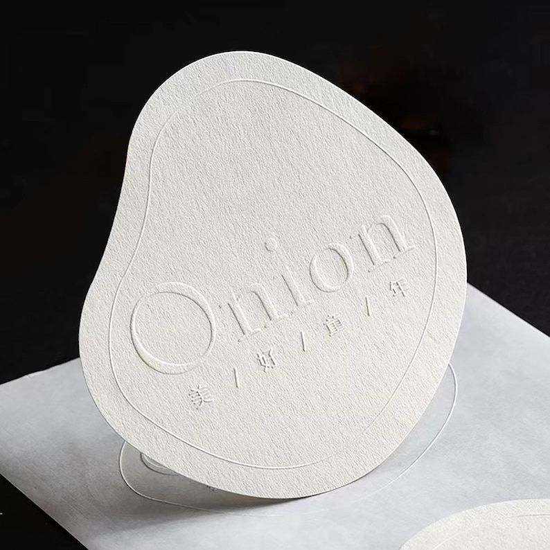 Puede incluir: Una tarjeta de papel blanco texturizado con la palabra "Onion" en relieve en el centro. La tarjeta tiene una forma org&aacute;nica &uacute;nica con bordes redondeados y elementos de dise&ntilde;o y texto en relieve.