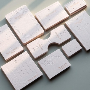 May include: A collection of off-white earring display cards with embossed designs and the text "NEW FASHION" and "Fashion Studio". Some cards have oval frames, others feature leaf patterns, and some have text that reads "Open a new day and it's most beautiful of you."