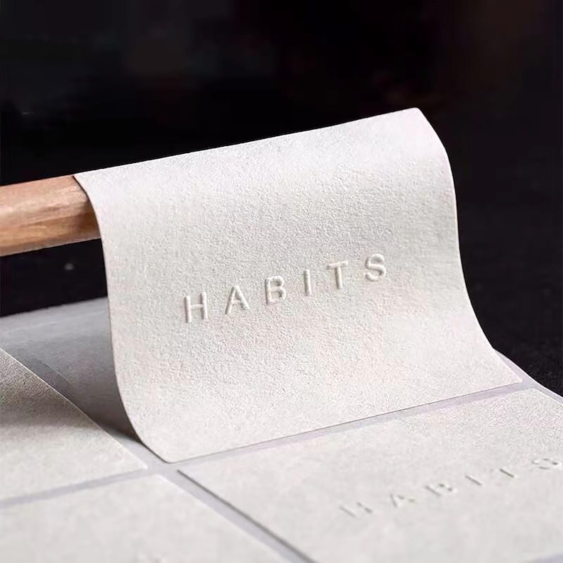 Puede incluir: Primer plano de una pila de hojas de papel blanco texturizado con la palabra "HABITS" en relieve en la hoja superior. El papel est&aacute; parcialmente despegado, revelando la palabra en las hojas subyacentes. Se ve una clavija de madera.