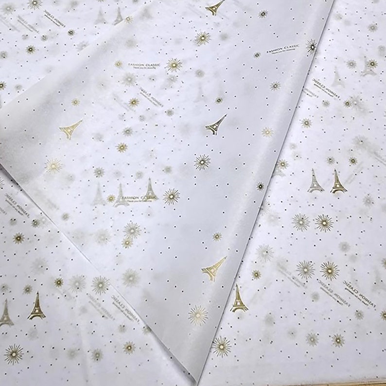 Może przedstawiać: Biały papier jedwabny z powtarzającym się wzorem złotych wież Eiffla i gwiazd. Na papierze nadrukowany jest tekst "FASHION CLASSIC".