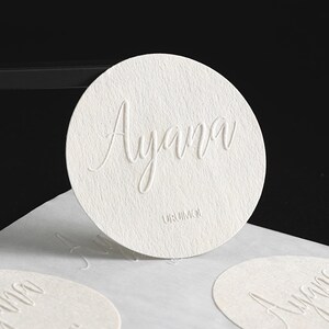 Puede incluir: Primer plano de un posavasos circular blanco y texturizado con el nombre "Ayana" en relieve en fuente cursiva. La palabra "URUMOI" est&aacute; impresa debajo. Se ven posavasos adicionales en primer plano.