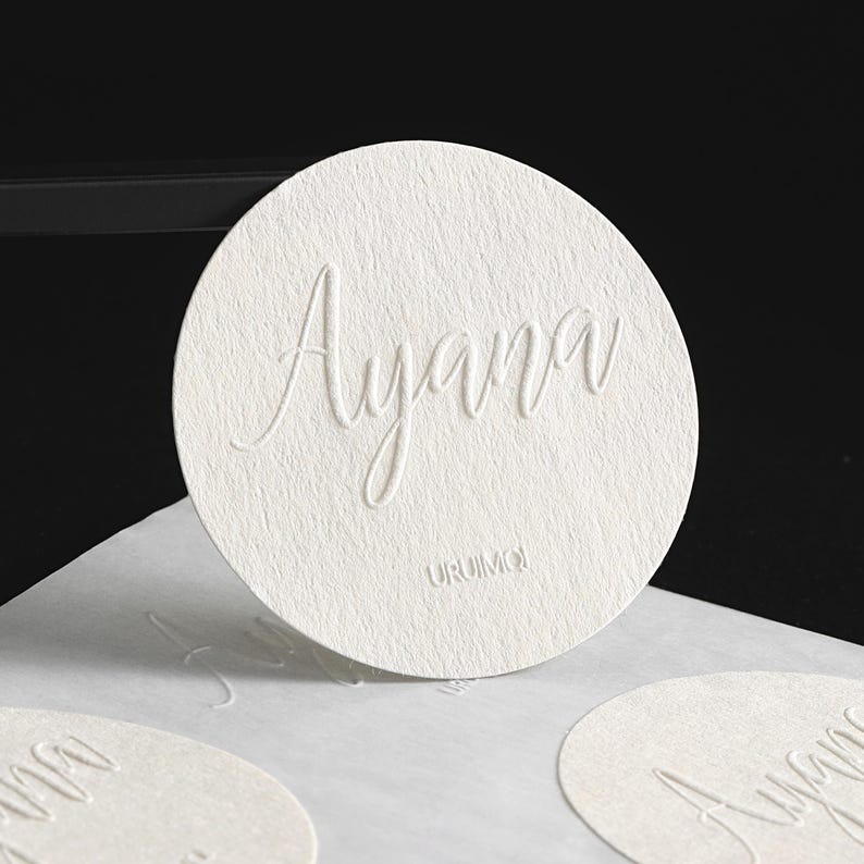 Puede incluir: Primer plano de un posavasos circular blanco y texturizado con el nombre "Ayana" en relieve en fuente cursiva. La palabra "URUMOI" est&aacute; impresa debajo. Se ven posavasos adicionales en primer plano.