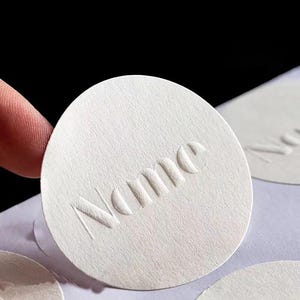 Puede incluir: Primer plano de una pegatina circular blanca y texturizada con la palabra "Name" en relieve. La pegatina est&aacute; sujeta por un dedo. Otras pegatinas similares son visibles en el fondo.