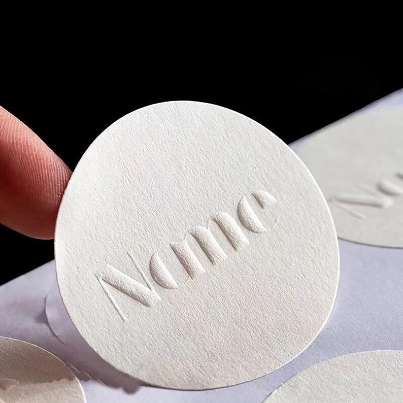 Puede incluir: Primer plano de una pegatina circular blanca y texturizada con la palabra "Name" en relieve. La pegatina est&aacute; sujeta por un dedo. Otras pegatinas similares son visibles en el fondo.