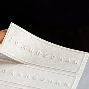 Puede incluir: Primer plano de varias etiquetas rectangulares blanquecinas con el nombre "JOANNECHAN" en relieve en may&uacute;sculas. Las etiquetas tienen una superficie texturizada y un borde sutil. El fondo es oscuro.