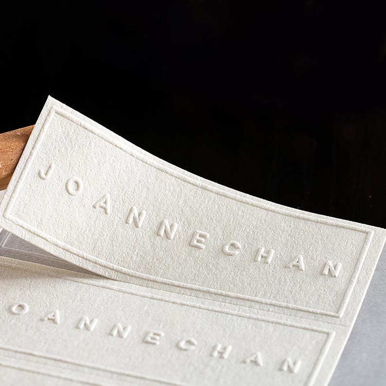 Puede incluir: Primer plano de varias etiquetas rectangulares blanquecinas con el nombre "JOANNECHAN" en relieve en may&uacute;sculas. Las etiquetas tienen una superficie texturizada y un borde sutil. El fondo es oscuro.