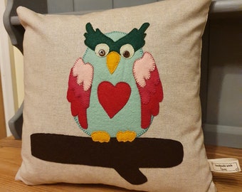May include: A beige linen cushion with a felt appliqué of an owl holding a red heart. The owl is green, pink, and yellow. The owl is sitting on a brown felt branch. The cushion has a tag that says "handmade with love".