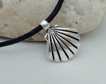 May include: A silver-colored seashell pendant on a black cord necklace. The pendant is detailed with radiating lines and a scalloped edge. The necklace is displayed against a white background and a light-colored rock.