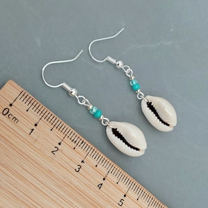 May include: A pair of silver earrings with turquoise beads and white cowrie shell charms. The earrings are shown against a wooden ruler with centimeter markings.