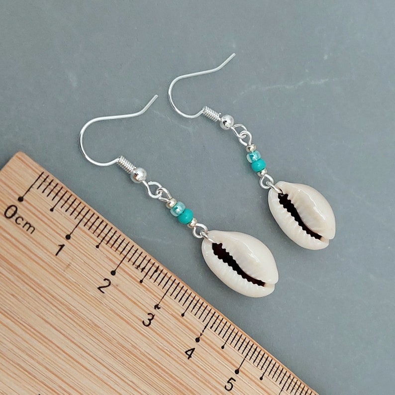 May include: A pair of silver earrings with turquoise beads and white cowrie shell charms. The earrings are shown against a wooden ruler with centimeter markings.