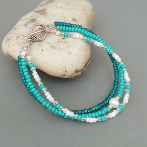 May include: A three-strand beaded bracelet featuring turquoise, white, and silver beads. The bracelet is secured with a toggle clasp and includes a pearl. The beads are small and evenly spaced, creating a delicate design.