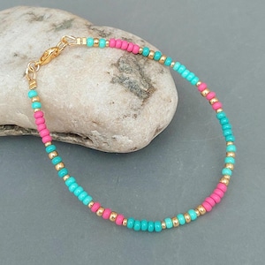 May include: A beaded bracelet with alternating sections of pink and turquoise beads, and gold-coloured beads. The bracelet is secured with a gold-toned clasp. The jewellery is displayed on a light grey stone.