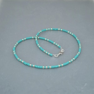 May include: A beaded necklace featuring alternating small turquoise blue and metallic silver beads arranged in a continuous circular pattern. The necklace is secured with a silver-toned metal lobster claw clasp and a small jump ring connector.