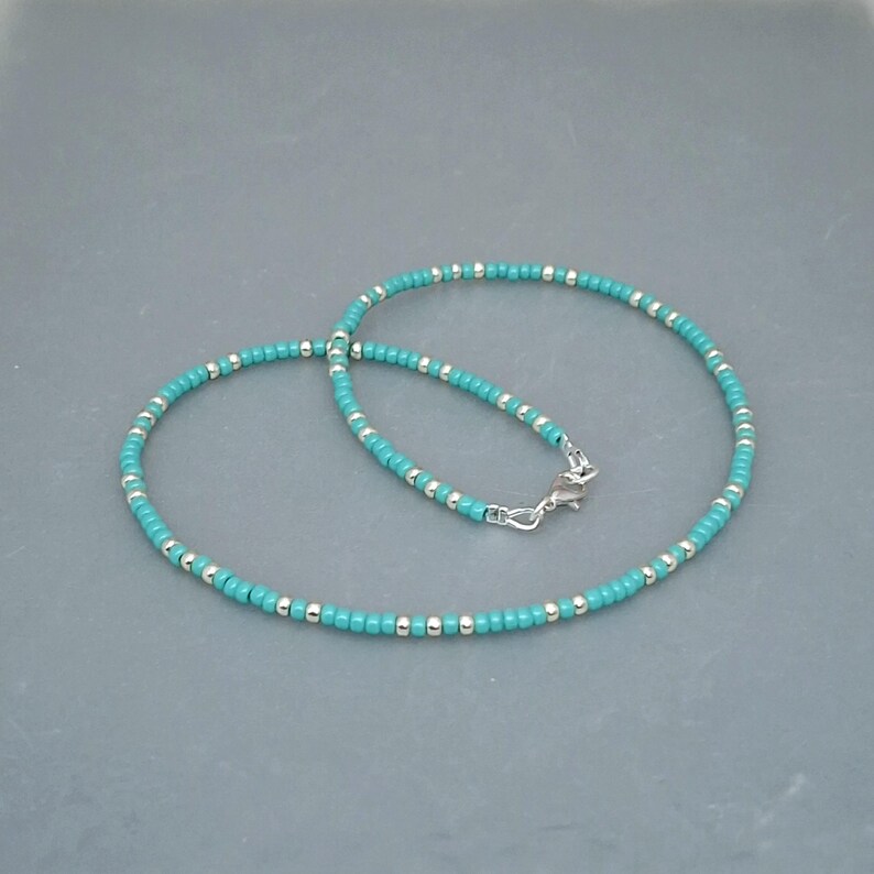 May include: A beaded necklace featuring alternating small turquoise blue and metallic silver beads arranged in a continuous circular pattern. The necklace is secured with a silver-toned metal lobster claw clasp and a small jump ring connector.