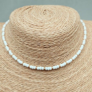 Peut inclure: Un collier ras de cou en perles blanches avec des accents dor&eacute;s. Le collier est sur un mannequin en paille.