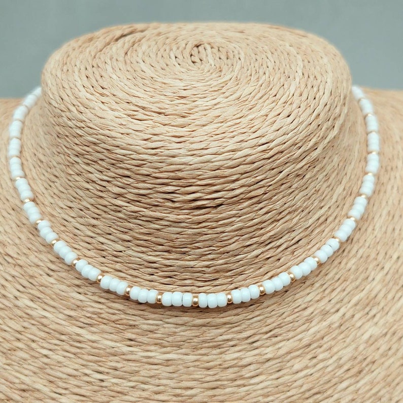 Peut inclure: Un collier ras de cou en perles blanches avec des accents dor&eacute;s. Le collier est sur un mannequin en paille.