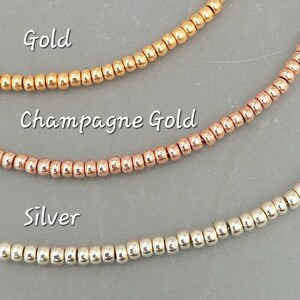 May include: Three strands of small, round beads in gold, champagne gold, and silver. The beads are arranged horizontally, with the gold beads at the top, the champagne gold beads in the middle, and the silver beads at the bottom. The words "Gold", "Champagne Gold", and "Silver" are written below each strand of beads.