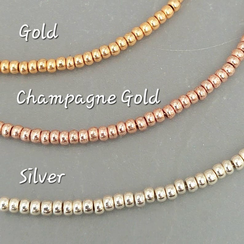 May include: Three strands of small, round beads in gold, champagne gold, and silver. The beads are arranged horizontally, with the gold beads at the top, the champagne gold beads in the middle, and the silver beads at the bottom. The words "Gold", "Champagne Gold", and "Silver" are written below each strand of beads.