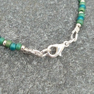 May include: A close-up of a silver clasp on a green and silver beaded bracelet.