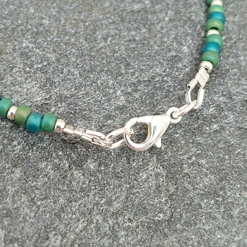May include: A close-up of a silver clasp on a green and silver beaded bracelet.
