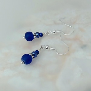 May include: A pair of dangle earrings featuring a dark blue glass bead and silver beads. The earrings have silver hooks.