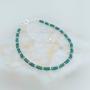 May include: A delicate silver chain bracelet with green and blue beads. The beads are small and round, and they are arranged in a simple pattern.