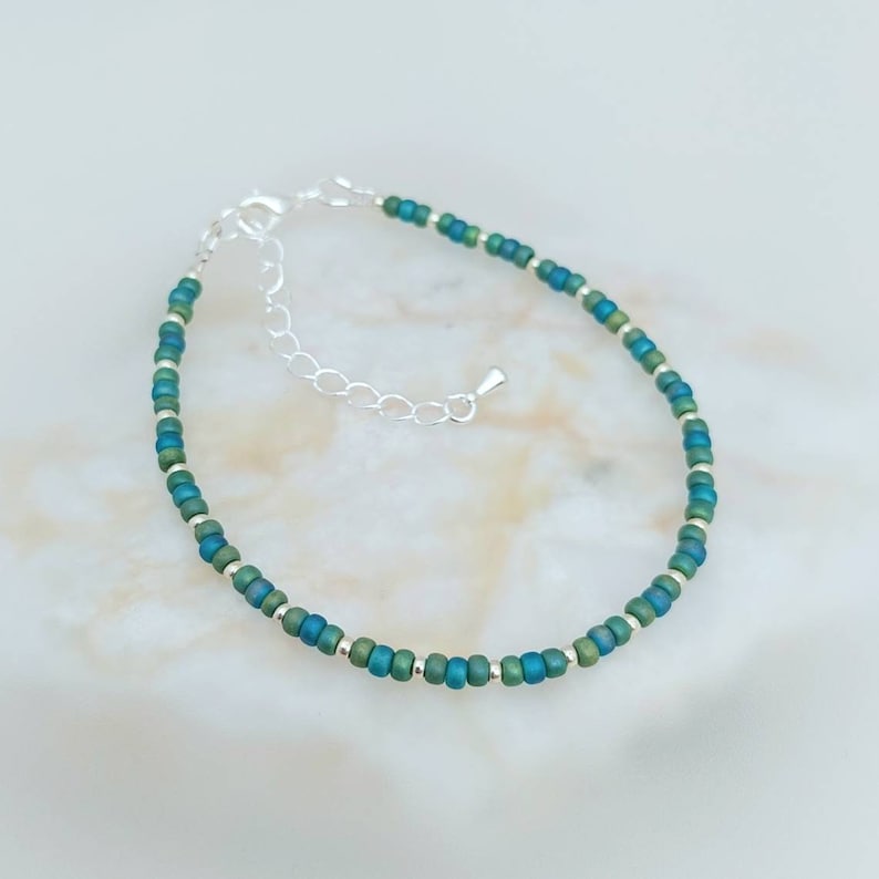 May include: A delicate silver chain bracelet with green and blue beads. The beads are small and round, and they are arranged in a simple pattern.