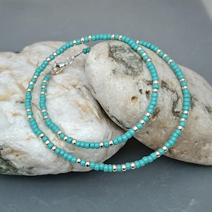 May include: A delicate beaded necklace or bracelet featuring a repeating pattern of small, round turquoise-colored beads interspersed with tiny, reflective silver-toned metallic beads. The jewelry piece includes a silver-toned metal lobster claw clasp.