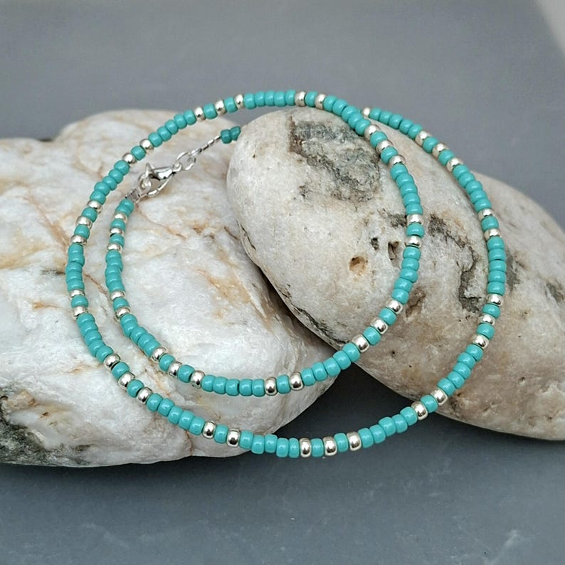 May include: A delicate beaded necklace or bracelet featuring a repeating pattern of small, round turquoise-colored beads interspersed with tiny, reflective silver-toned metallic beads. The jewelry piece includes a silver-toned metal lobster claw clasp.