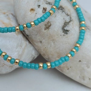 May include: A beaded necklace or bracelet featuring a repeating pattern of small, round, opaque turquoise blue beads interspersed with single, metallic gold-toned spherical beads arranged in a continuous, circular strand design.