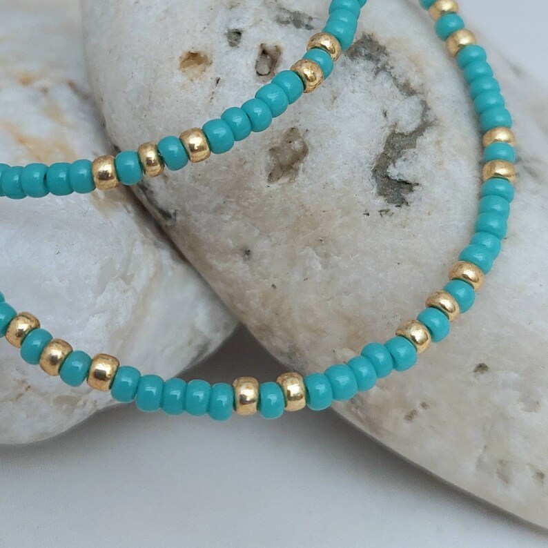 May include: A beaded necklace or bracelet featuring a repeating pattern of small, round, opaque turquoise blue beads interspersed with single, metallic gold-toned spherical beads arranged in a continuous, circular strand design.