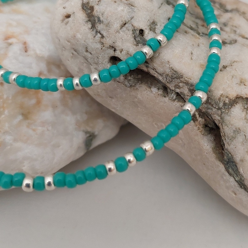 May include: A beaded necklace or bracelet featuring a repeating pattern of small, round, vibrant turquoise blue beads interspersed with single, small, metallic silver-colored spacer beads, creating a simple and uniform design throughout the strand.
