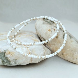 Peut inclure: Un collier de perles blanches avec des perles argent&eacute;es entrem&ecirc;l&eacute;es. Le collier est pos&eacute; sur deux rochers lisses et gris.