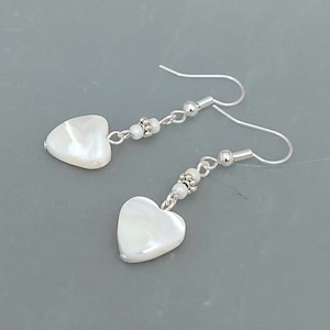 May include: A pair of silver earrings with white heart-shaped pendants. The earrings are made with small silver beads and have a simple design.