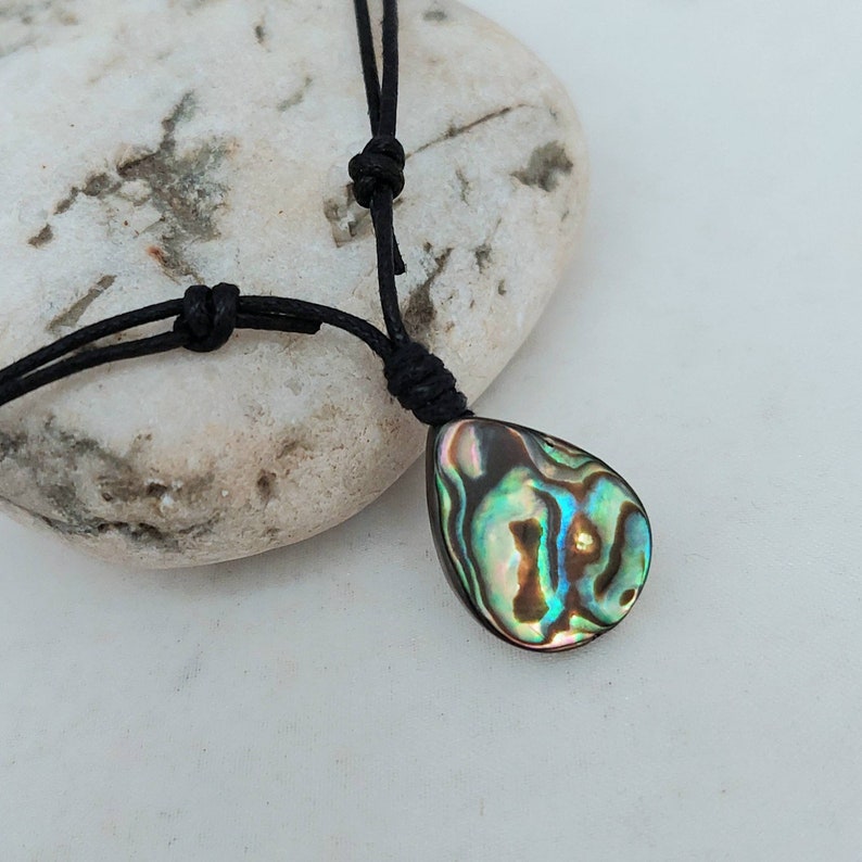 May include: A black cord necklace with a teardrop-shaped abalone shell pendant. The shell has iridescent green, blue, and brown colors.