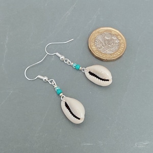 May include: A pair of silver earrings with turquoise beads and cowrie shell charms. The earrings are approximately the size of a British one pound coin.