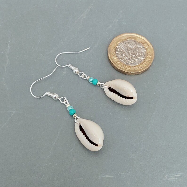 May include: A pair of silver earrings with turquoise beads and cowrie shell charms. The earrings are approximately the size of a British one pound coin.
