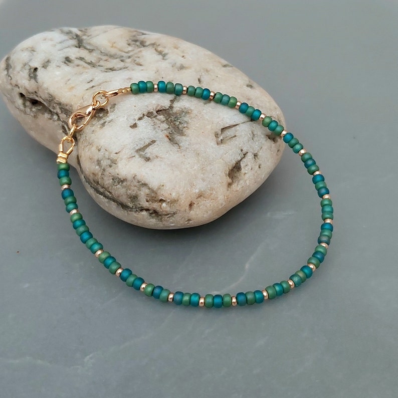 May include: A delicate bracelet made with small, iridescent green and gold beads. The bracelet has a gold clasp.