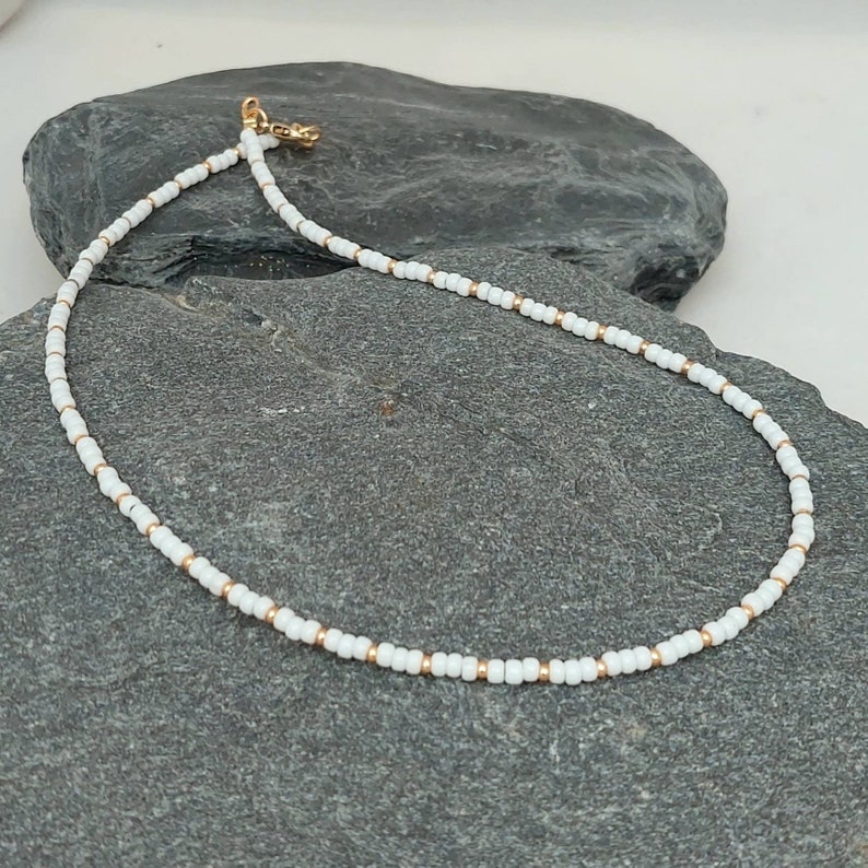 Peut inclure: Un collier d&eacute;licat en perles blanches avec des accents dor&eacute;s. Le collier est compos&eacute; de petites perles rondes et pr&eacute;sente un design simple et minimaliste.