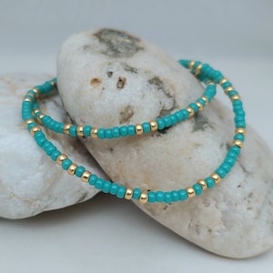 May include: A beaded bracelet featuring a repeating pattern of small, round, opaque turquoise beads and metallic gold-toned spacer beads. The circular jewelry piece is constructed with a flexible cord, showcasing a consistent alternating color design.