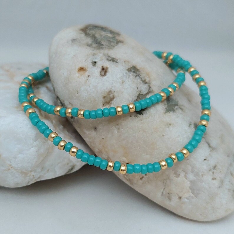 May include: A beaded bracelet featuring a repeating pattern of small, round, opaque turquoise beads and metallic gold-toned spacer beads. The circular jewelry piece is constructed with a flexible cord, showcasing a consistent alternating color design.