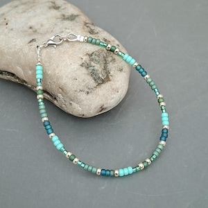 May include: A beaded bracelet featuring turquoise, teal, and green beads, with small silver-coloured beads. The bracelet is fastened with a silver-coloured clasp. The bracelet is shown on a light grey stone.