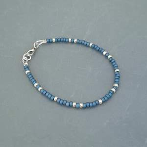 May include: A beaded bracelet featuring small matte slate blue seed beads interspersed with polished silver-toned metallic spacer beads. The bracelet is secured with a silver-toned metal lobster claw clasp and a small circular jump ring connector.