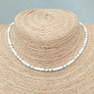 Peut inclure: Un collier ras de cou en perles blanches avec des accents argent&eacute;s. Le collier est sur un mannequin en paille.