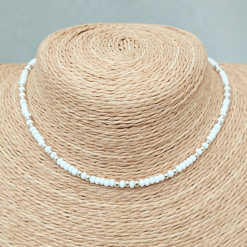 Peut inclure: Un collier ras de cou en perles blanches avec des accents argent&eacute;s. Le collier est sur un mannequin en paille.