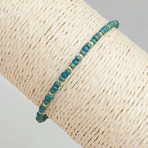 May include: A delicate bracelet made with small, iridescent green and silver beads. The bracelet is displayed on a beige, woven surface.