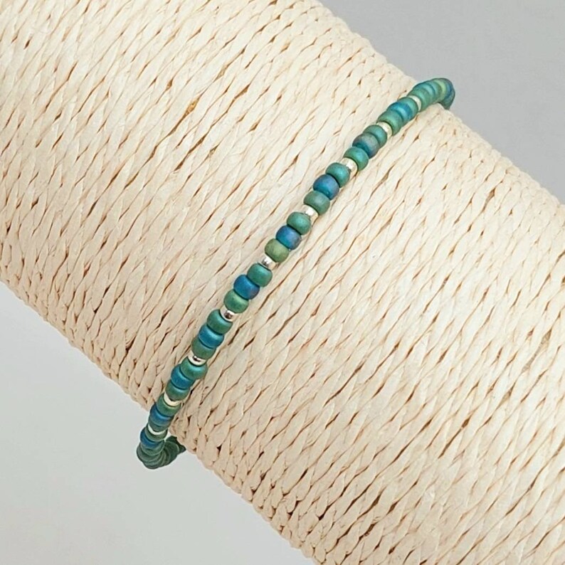 May include: A delicate bracelet made with small, iridescent green and silver beads. The bracelet is displayed on a beige, woven surface.