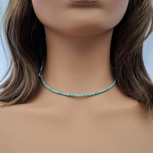 May include: A delicate choker necklace featuring a repeating pattern of small, round turquoise-colored beads interspersed with tiny, metallic silver-toned spacer beads, creating a simple and uniform circular design around the neck.